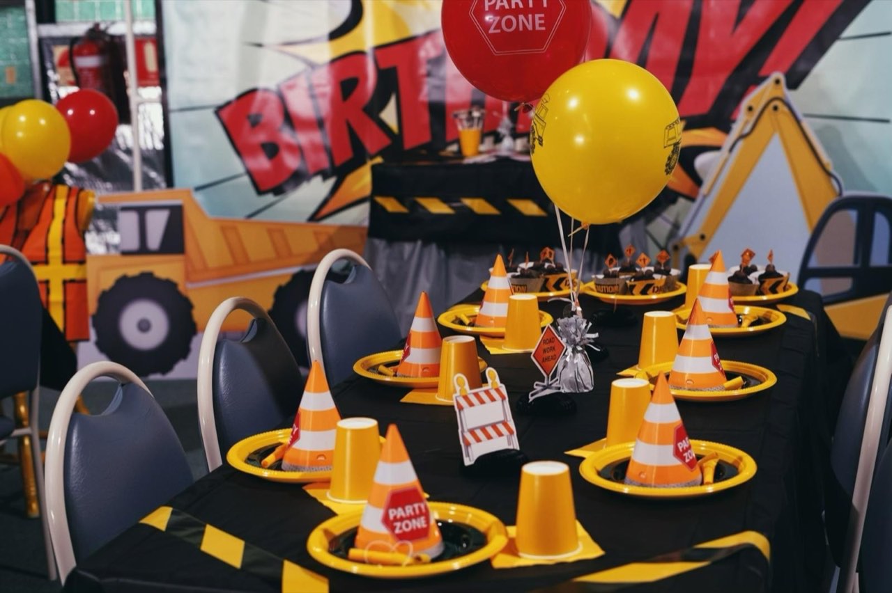 Construction-themed birthday party table with traffic-cone party hats, balloons, and excavator backdrop